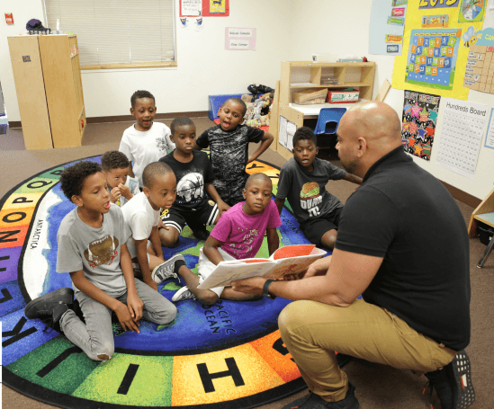 a group of young Black boys listens to a story being read by a Black, male teacher