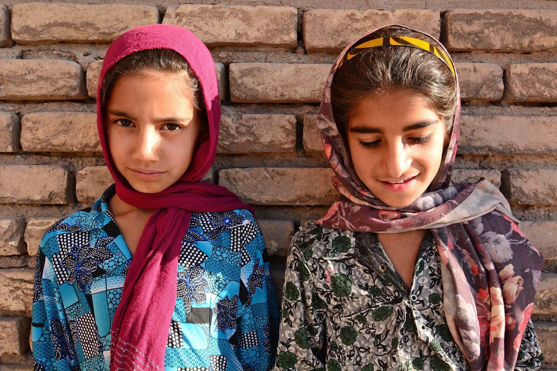 Two young Afghan girls stand against a brick wall