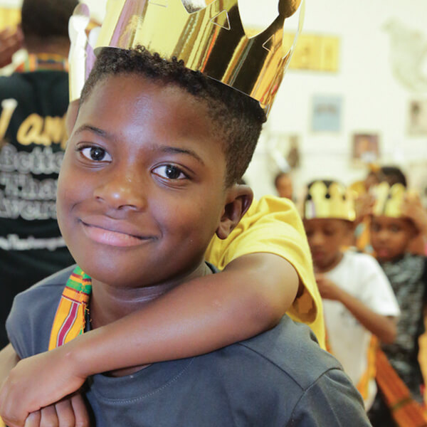 A young Black boy wearing a paper crown in a classroom full of his peers